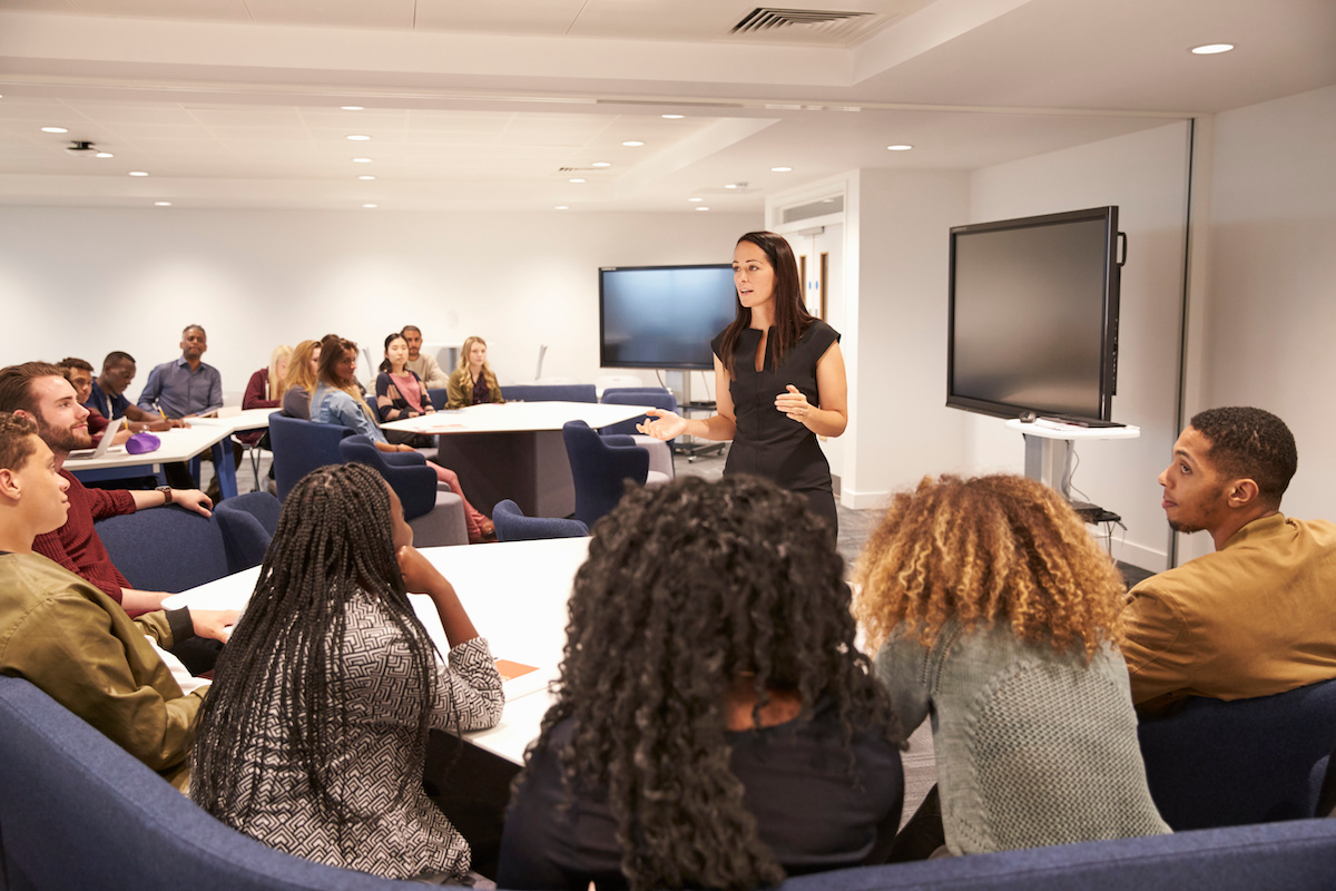 Woman stands at the front of the class speaking while students listen attentively.