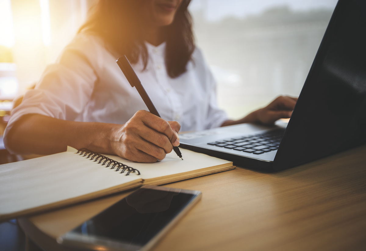 A young woman sits at a desk behind a computer taking notes.