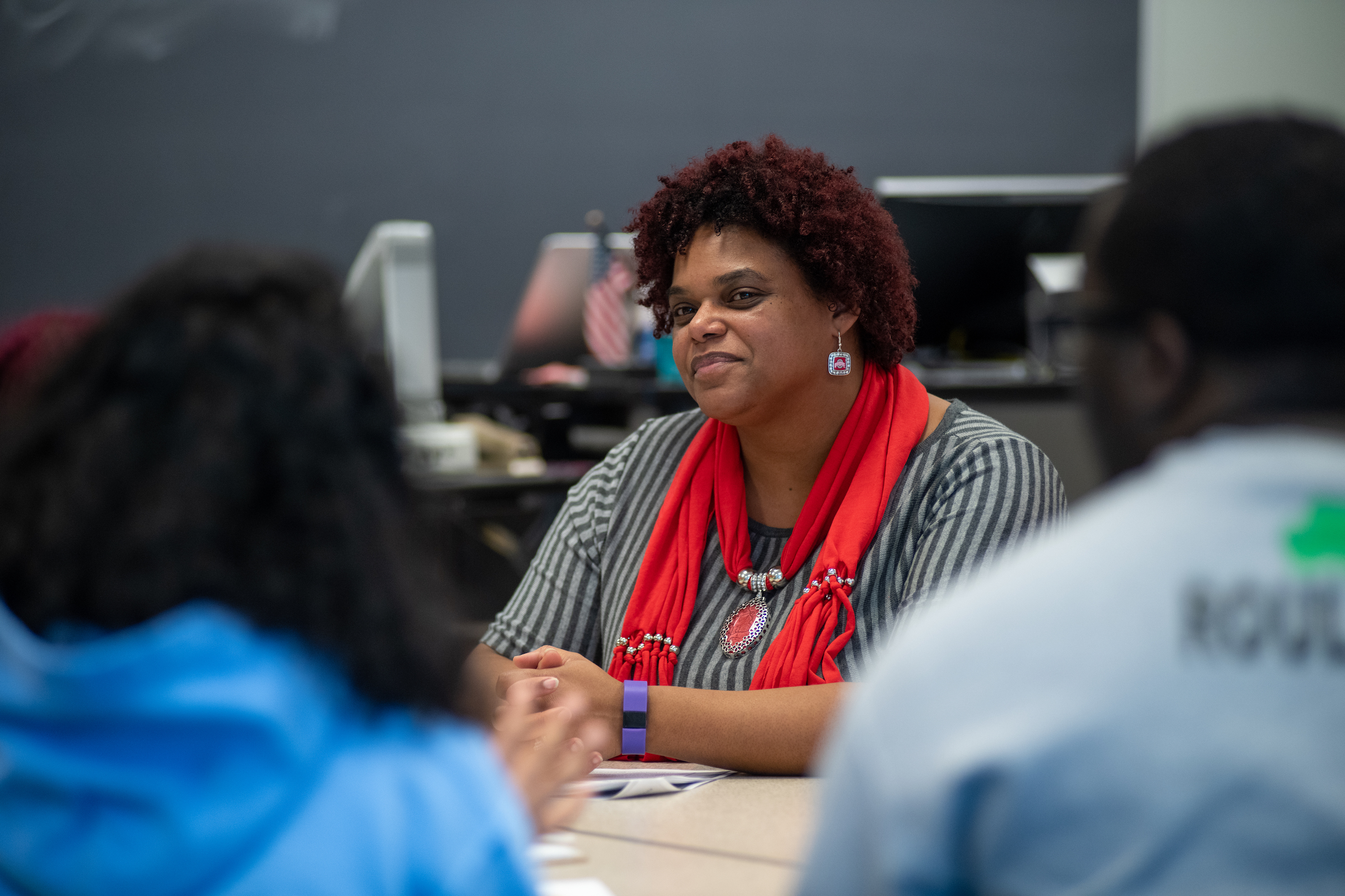 A professor sits in focus in front of two students in the foreground out of focus. The Faculty member is wearing a black and white blouse with a red scarf.