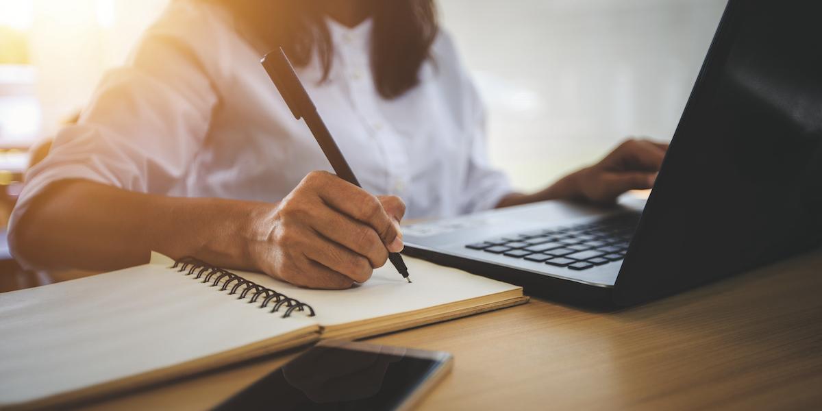 A young woman sits at a desk behind a computer taking notes.