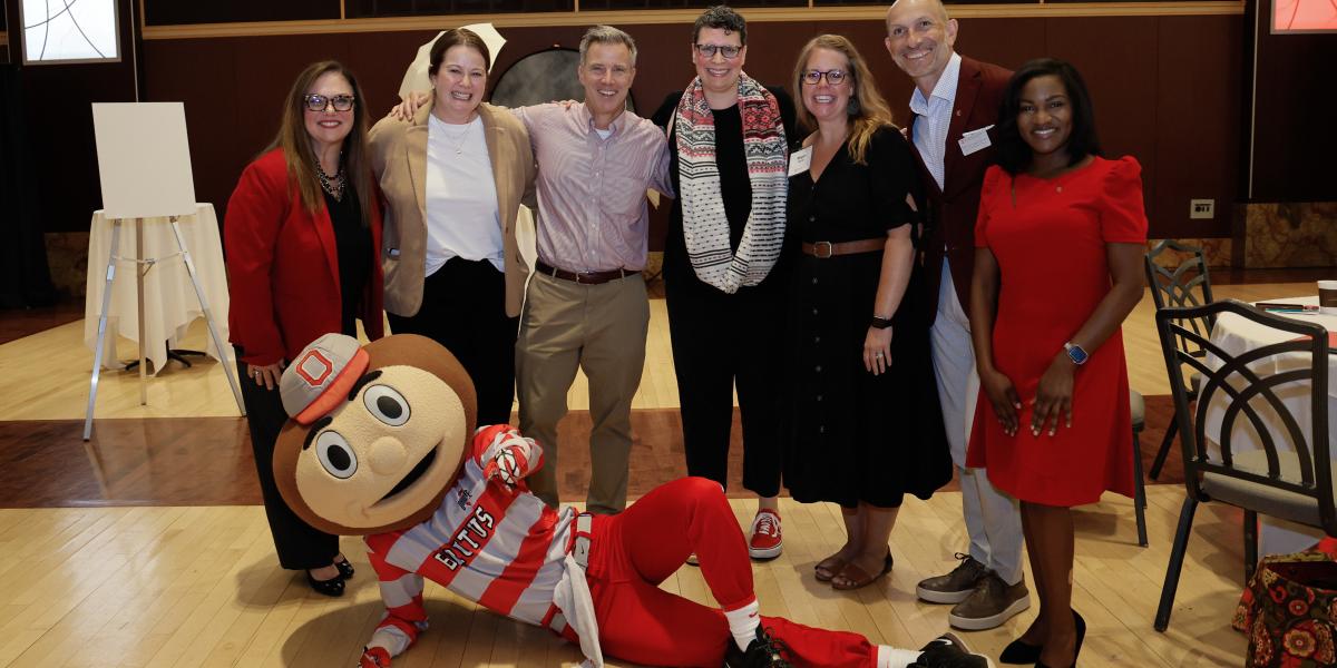 A group of smiling people stand behind Ohio State's mascot Brutus, who lies on his side on the floor.