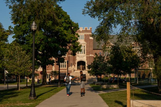 Two students walk on the oval under blue skies and lush trees.