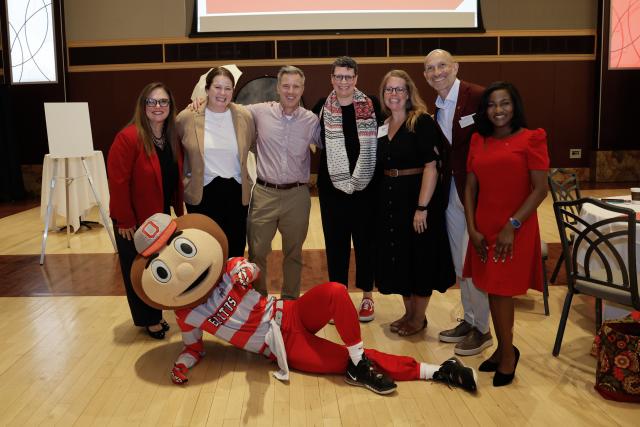 A group of smiling people stand behind Ohio State's mascot Brutus, who lies on his side on the floor.