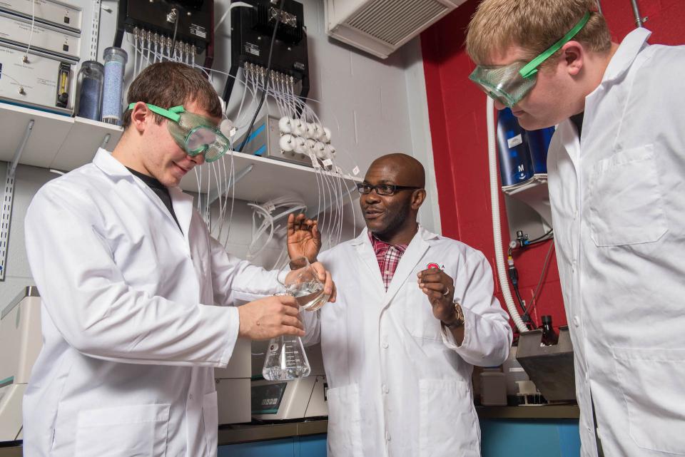 A clinical faculty instructor speaks with two students in a lab. All three wear lab coats and one holds a beaker.