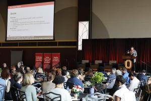 Picture of New Faculty Orientation shows an audience listening to Helen Malone speak from stage. To the left of the stage, a presentation is being projected on to a screen.